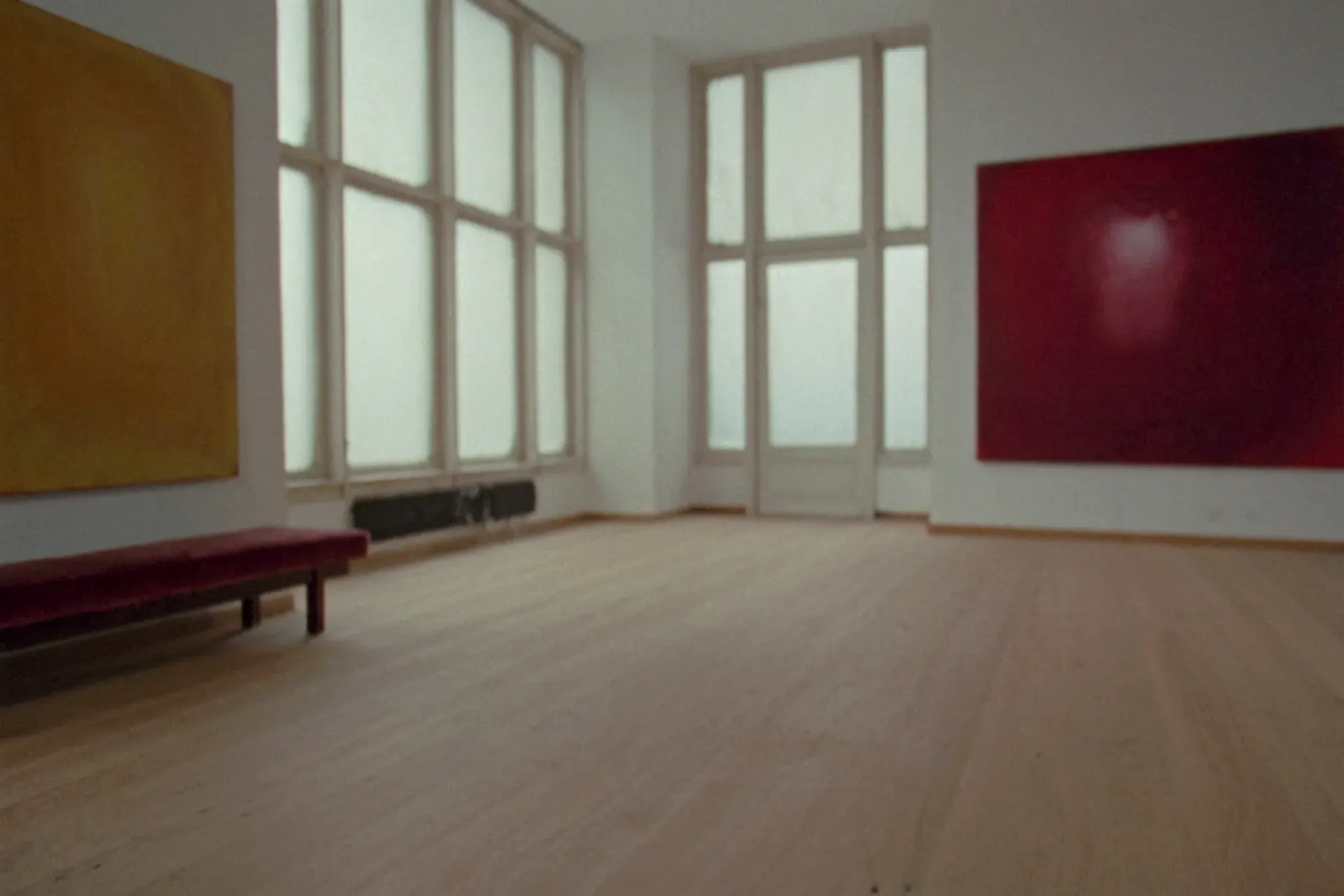 Lege galeriezaal met een geel en een donkerrood doek, hoge ramen en een fluwelen bank